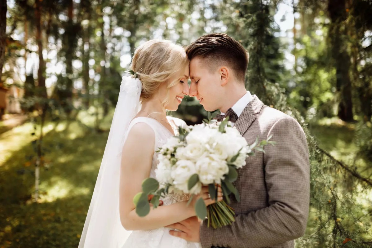 Un couple de jeunes mariés se tient front contre front dans un cadre boisé. La mariée porte une robe blanche élégante et un bouquet de fleurs blanches, tandis que le marié est en costume beige.